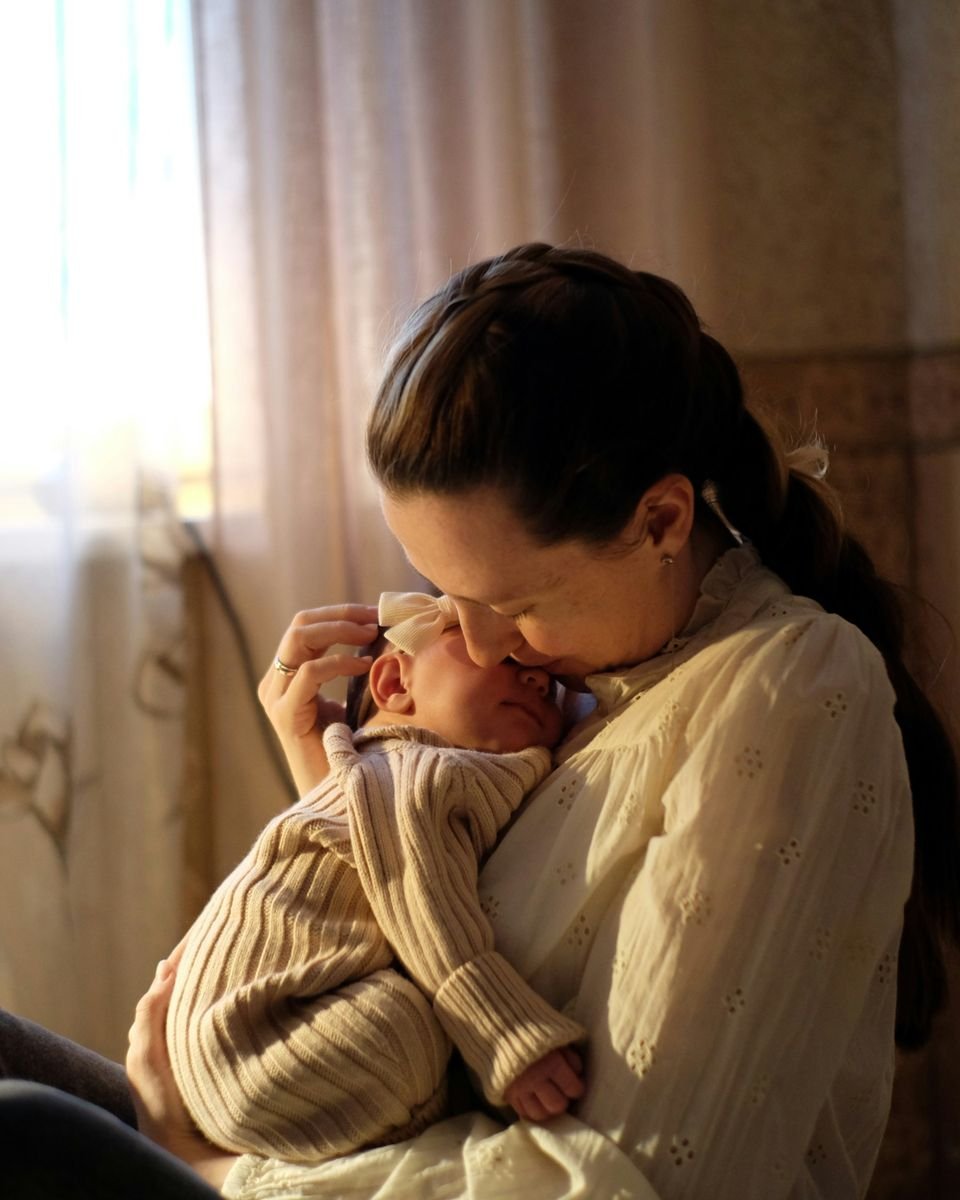 A mother holds her newborn close and kisses the baby's head, in soft light by a window.