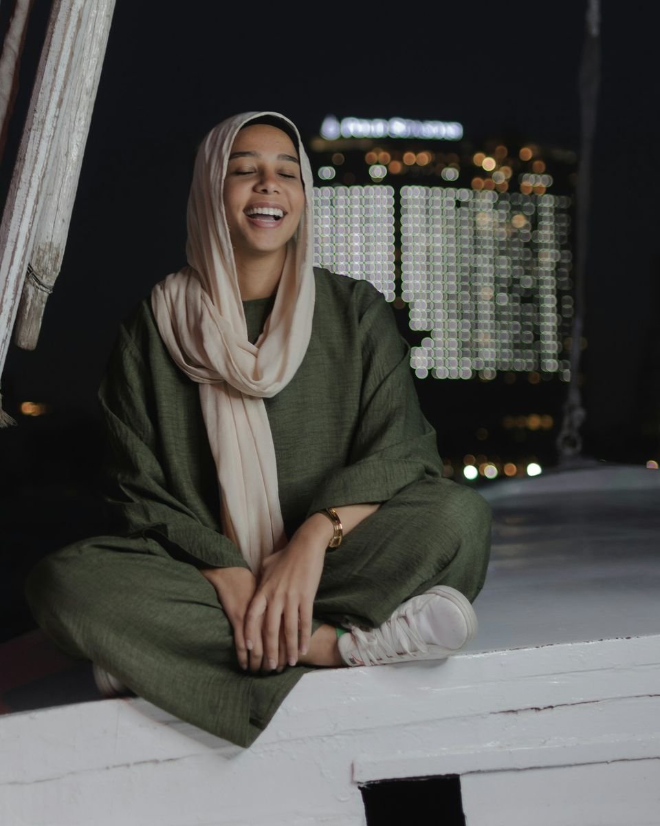 A young woman in a cream headscarf and olive-green clothes laughs with her eyes closed, at night, with city lights behind her.