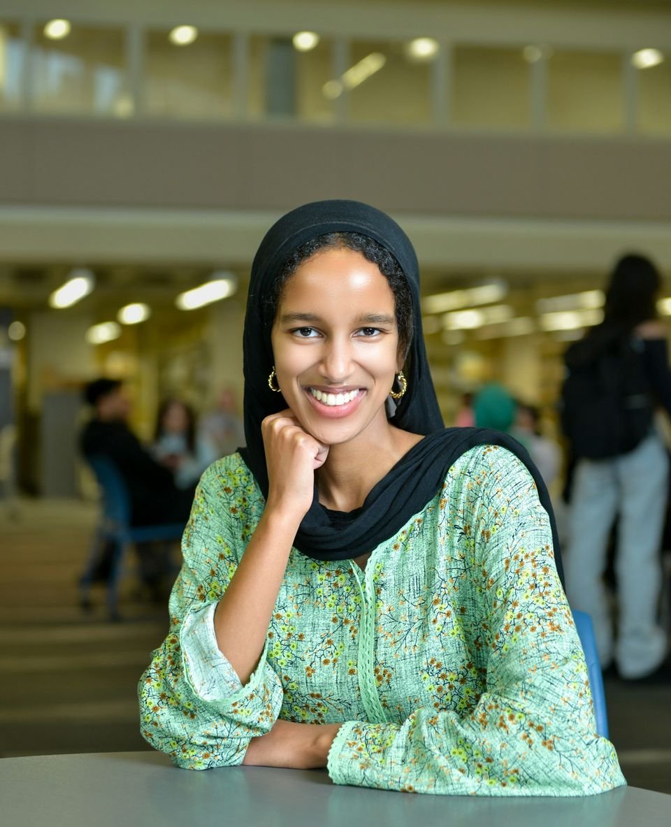 A young woman in a black headscarf and a green patterned top smiles, her chin resting on her hand, at a table in a library.